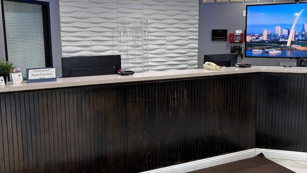 A reception desk with a cityscape on the monitor, a blue-lit ceiling, and a sign for key drop-off. - Travelodge by Wyndham St. Louis (St. Louis)