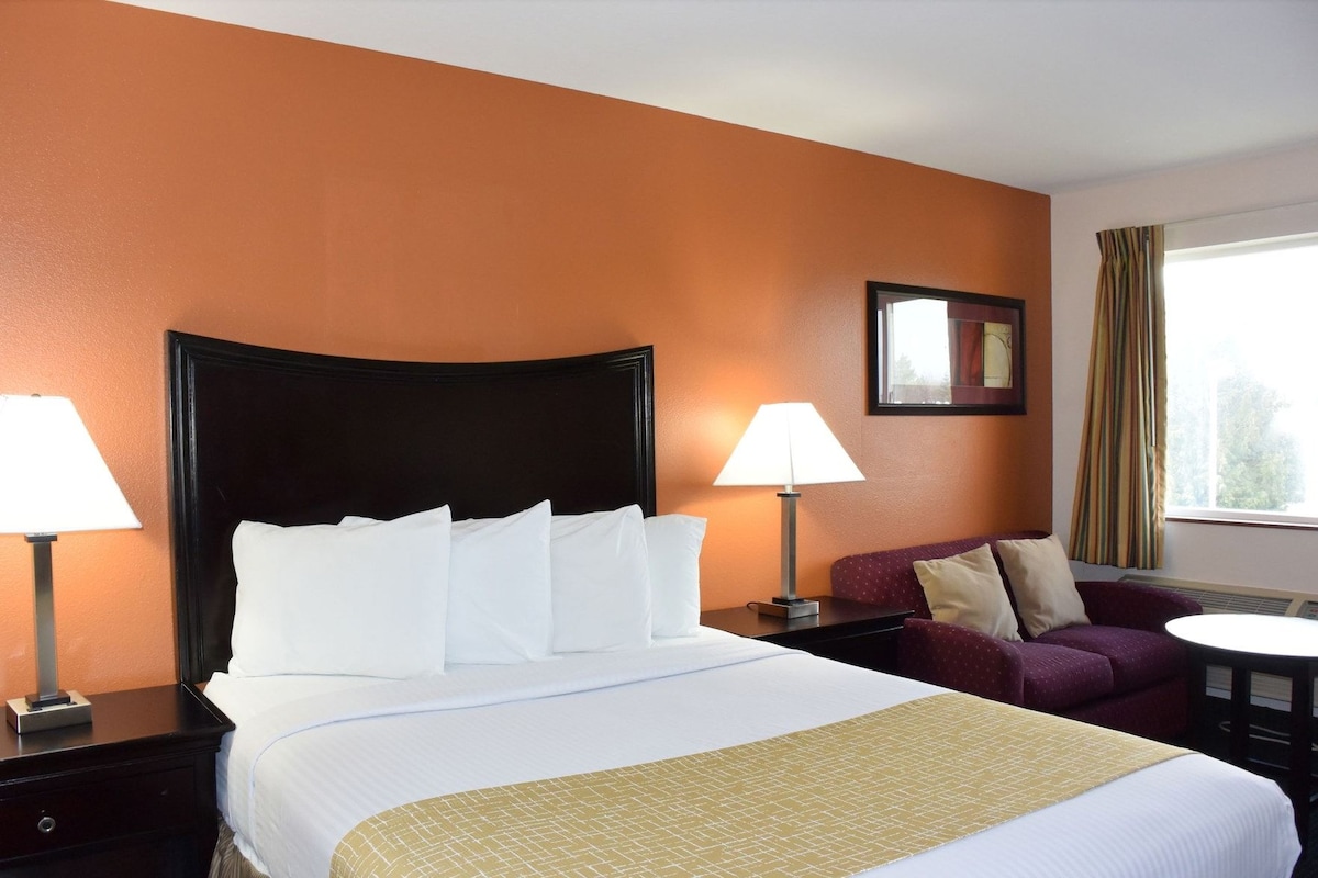 A hotel room with a large bed, two bedside lamps, a small table, a purple armchair, and a window with curtains.