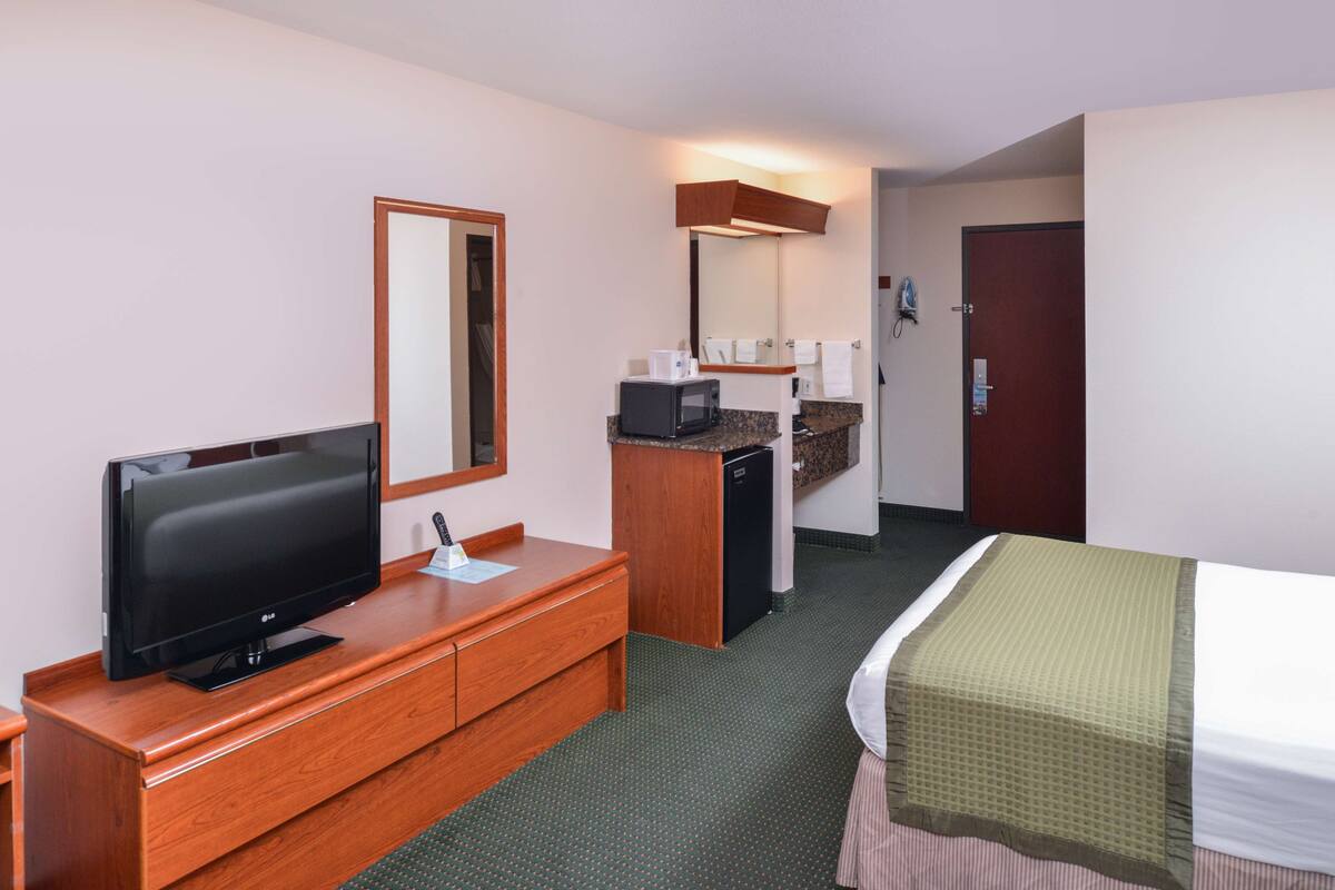 Ein Hotelzimmer mit Fernseher auf einer Holzkommode, einer kleinen Küchenzeile mit Mikrowelle und einem Bett mit grün-weiß gestreifter Bettdecke.