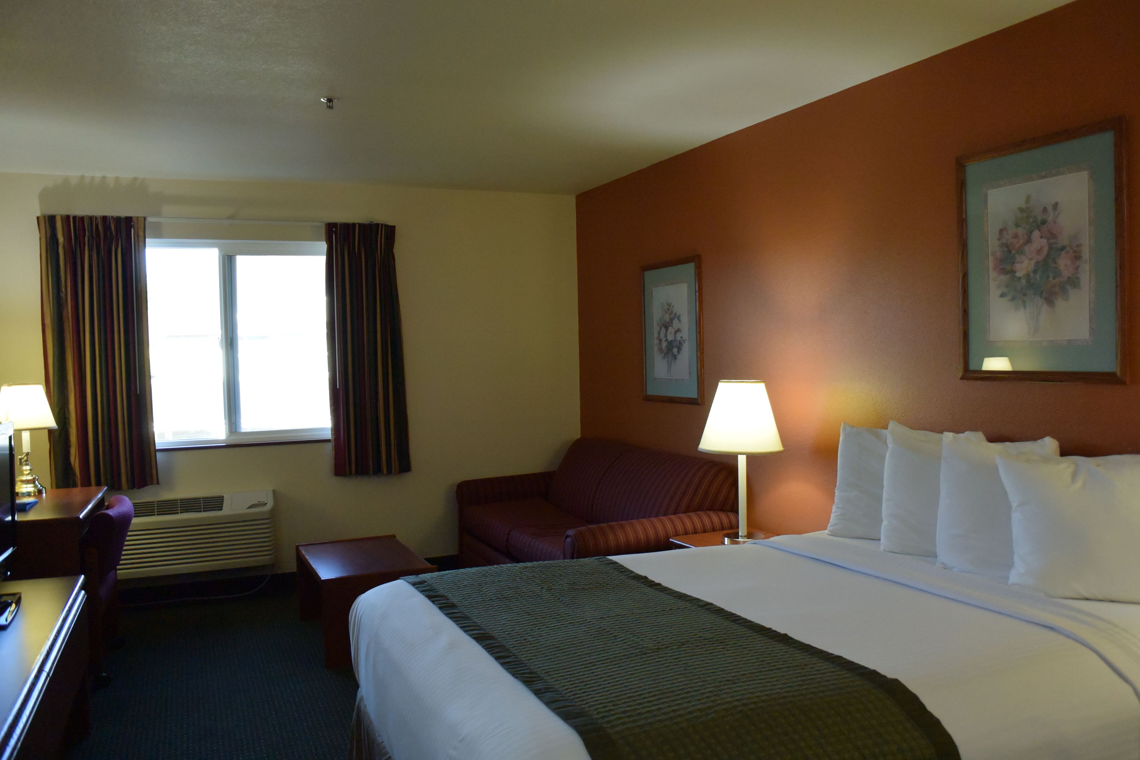 Ett hotellrum med en säng, ett skrivbord, en stol, en soffa, ett fönster med gardiner, en luftkonditionering, en lampa och en inramad tavla på väggen.