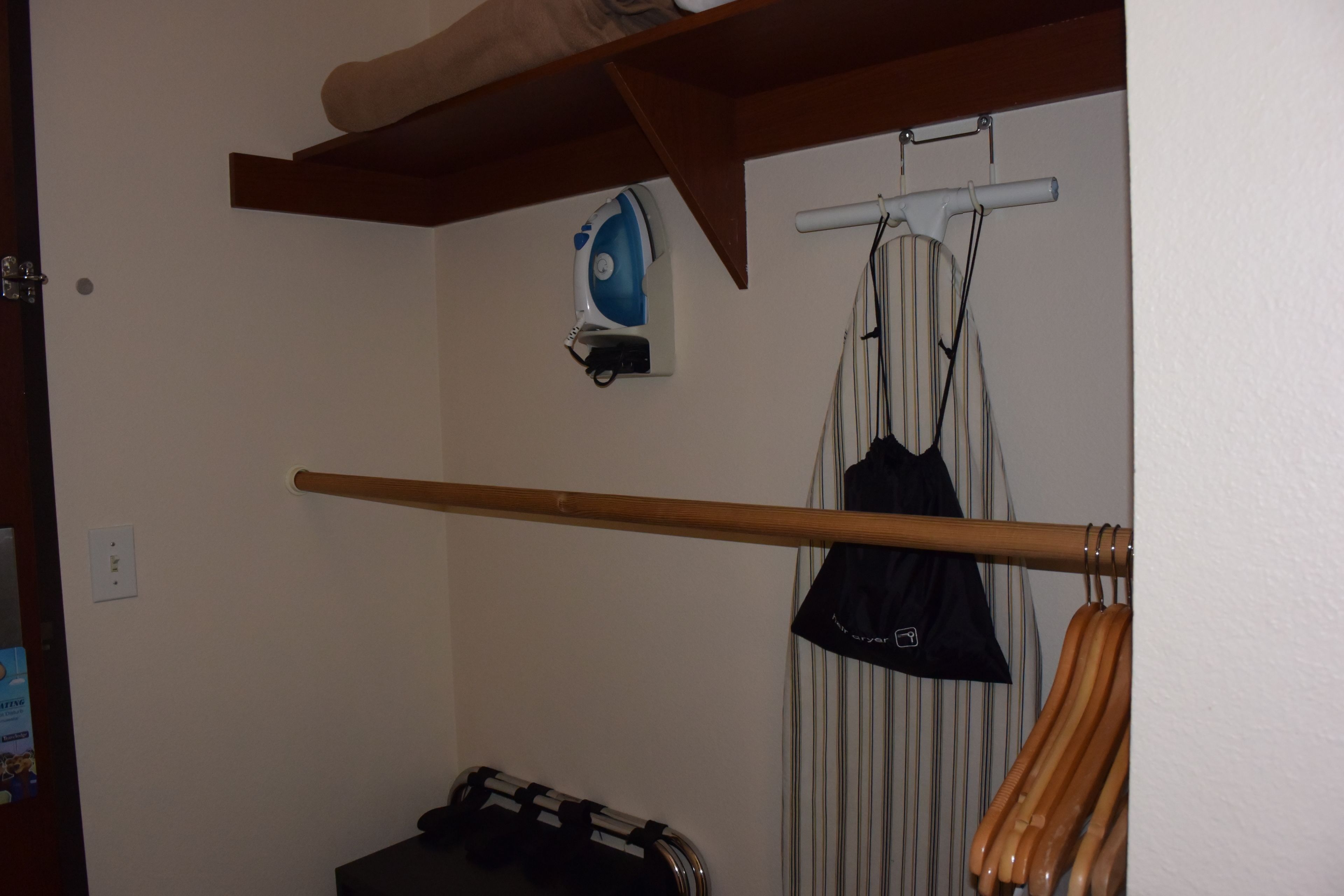 Ein Zimmer mit Holzbalkendecke, einer Kleiderstange, einem schwarzen Koffer und einem an der Wand befestigten Bügelbrett.