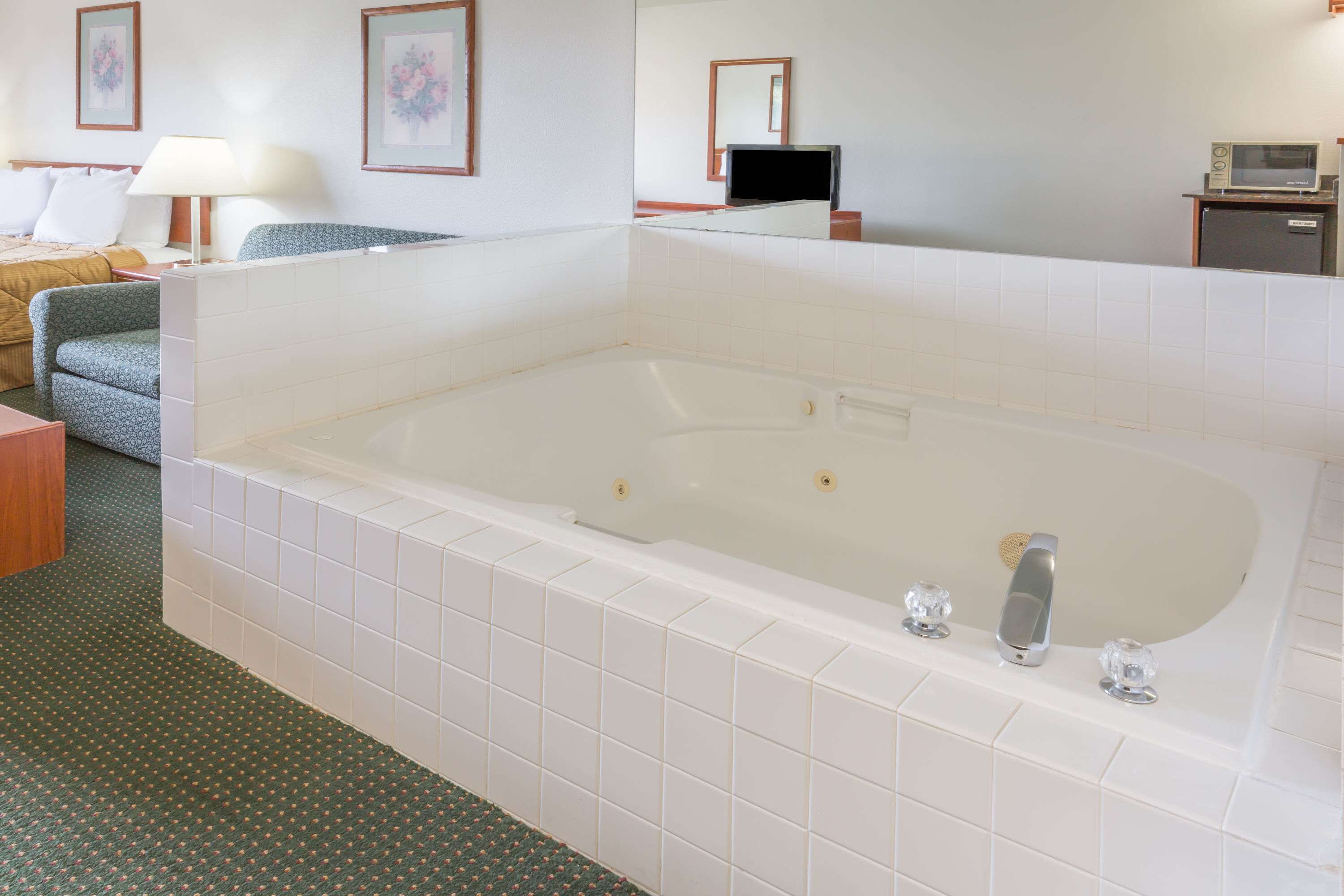 A hotel room with a large white bathtub, a green sofa, a wooden side table, a lamp, a television, and a microwave.