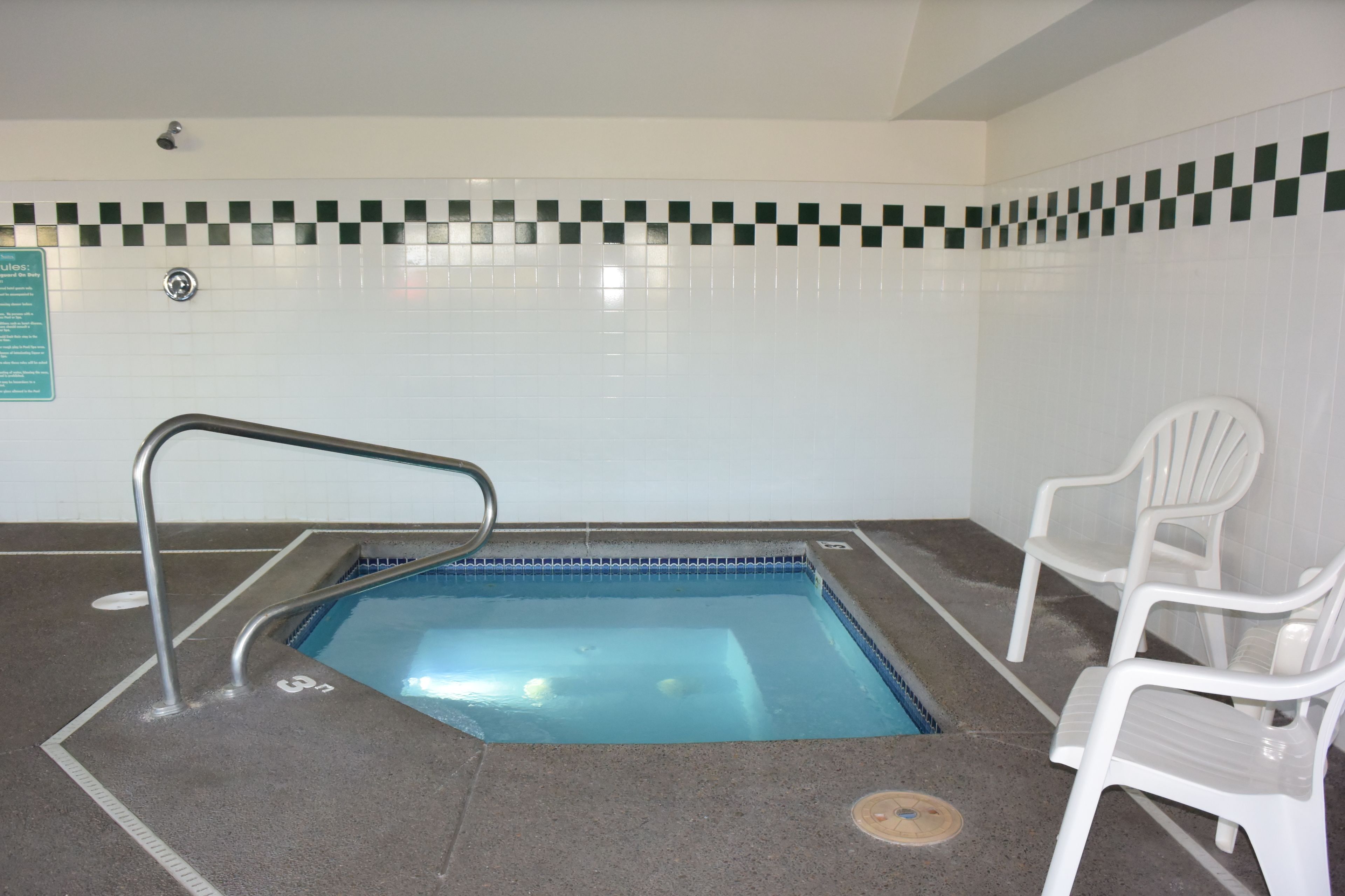 An indoor swimming pool with a tiled wall, a metal pool ladder, and two white plastic chairs.