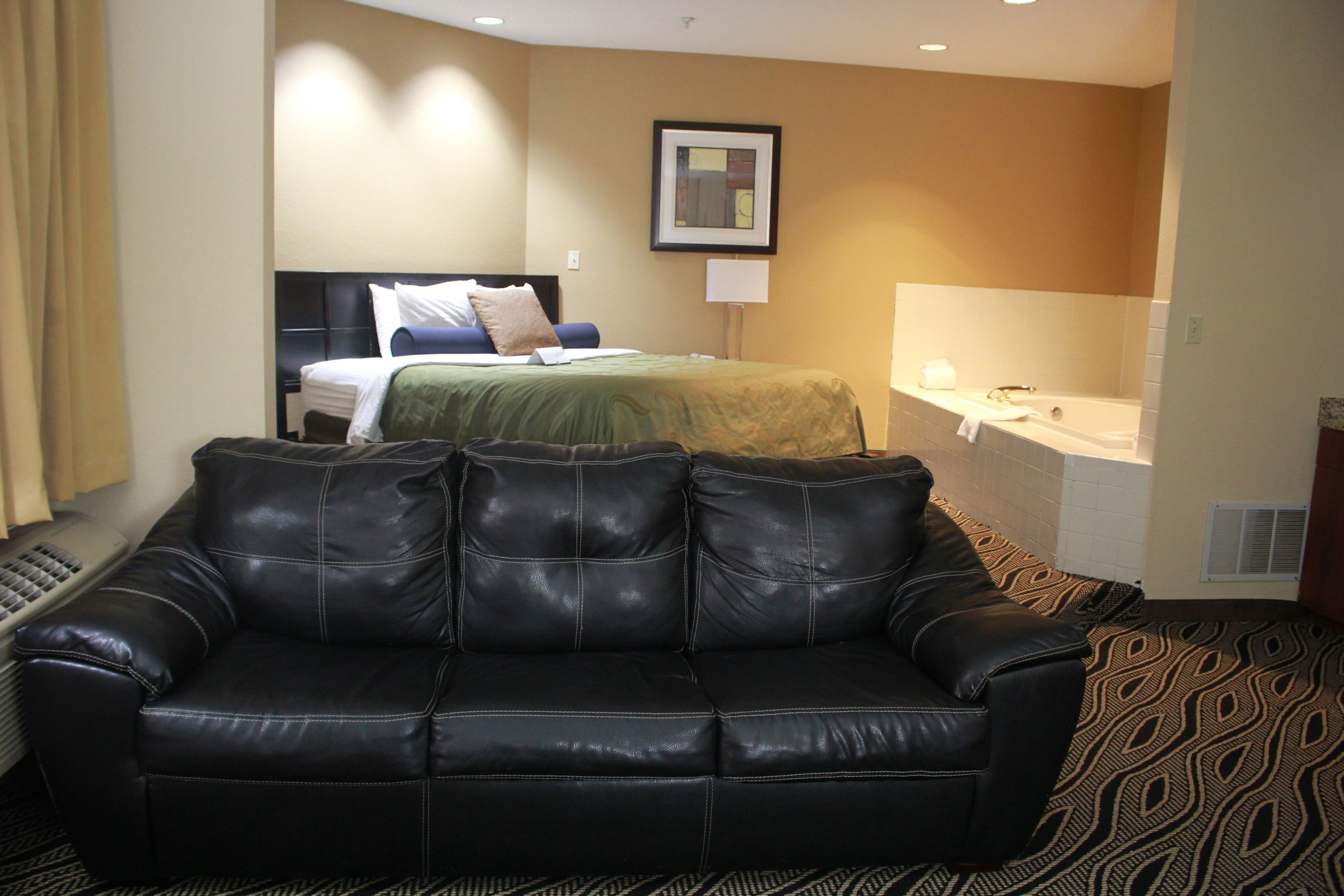 A hotel room with a black leather sofa, a bed with a green blanket, a framed picture on the wall, and a bathroom with a sink and mirror.