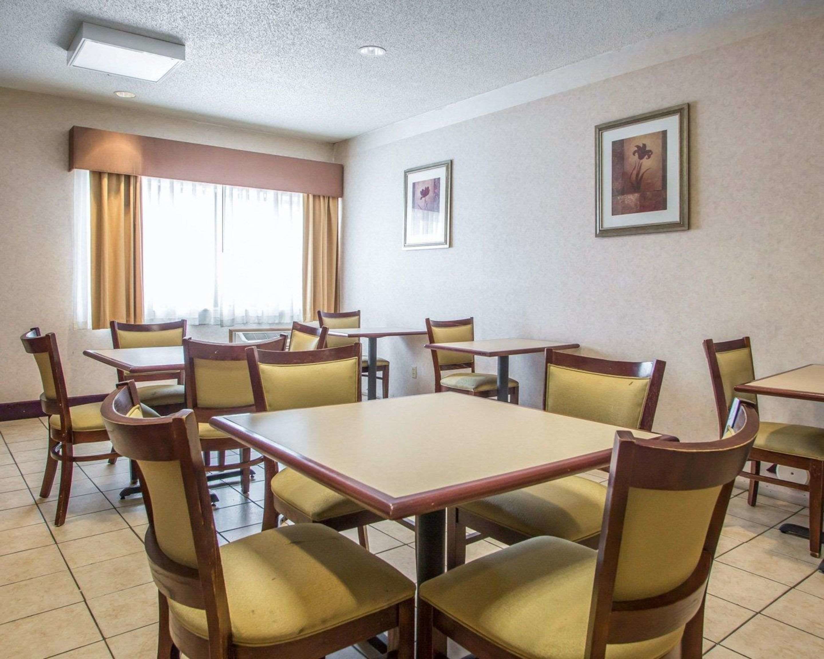 A hotel dining area with multiple tables and chairs, large windows with curtains, and framed artwork on the walls.