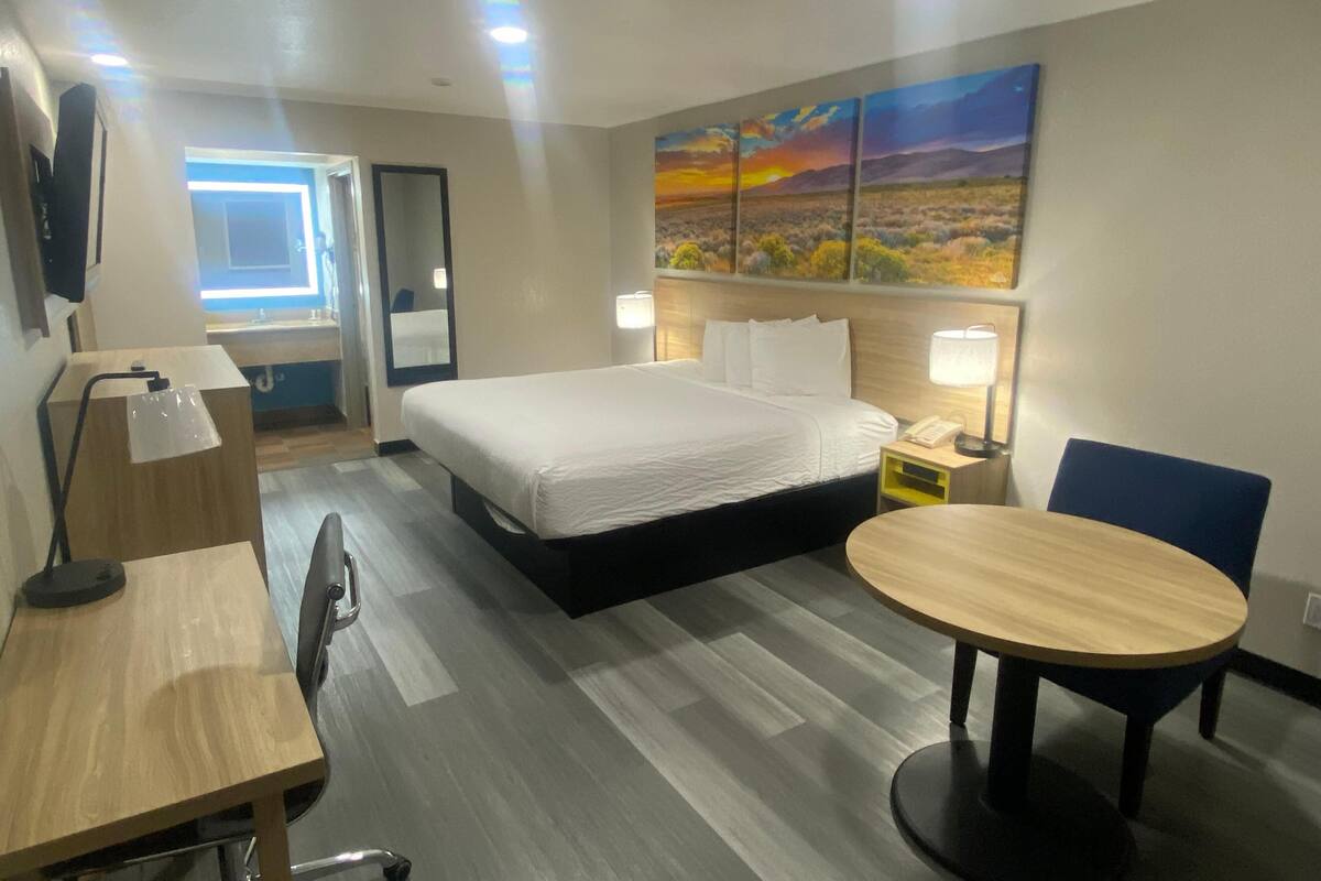 A hotel room with a bed, a desk, a chair, a round table, a nightstand, a lamp, a mirror, a TV, and a painting on the wall.
