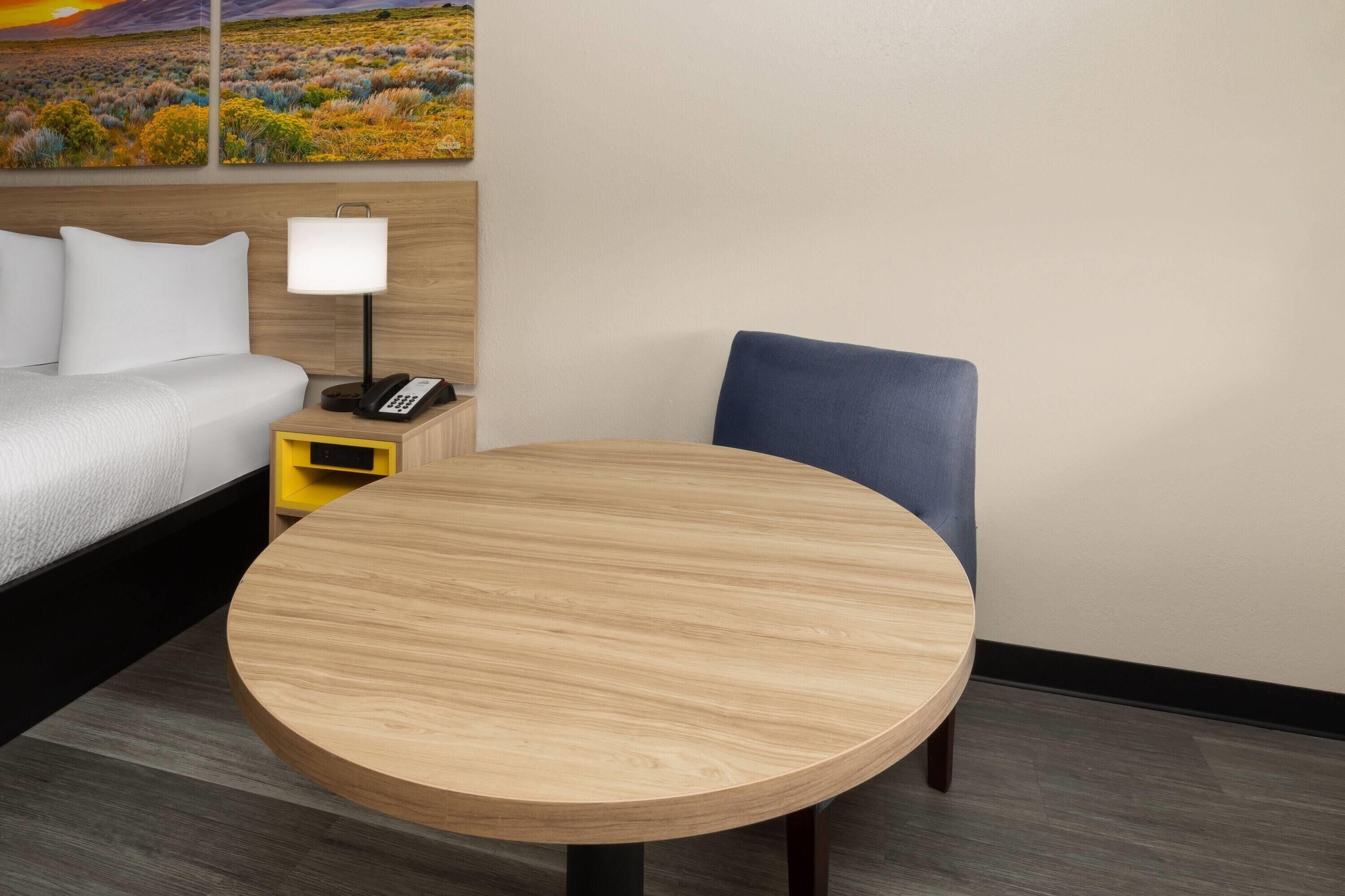 Habitación de hotel con una cama, una mesita de noche con lámpara, un teléfono, una mesa redonda de madera y una silla.