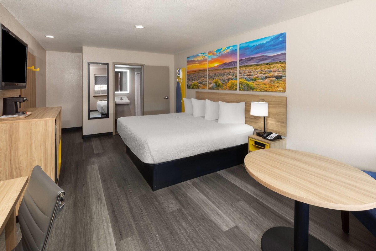 A hotel room with a bed, a desk, a chair, a small table, a TV, a bathroom, and a painting on the wall.