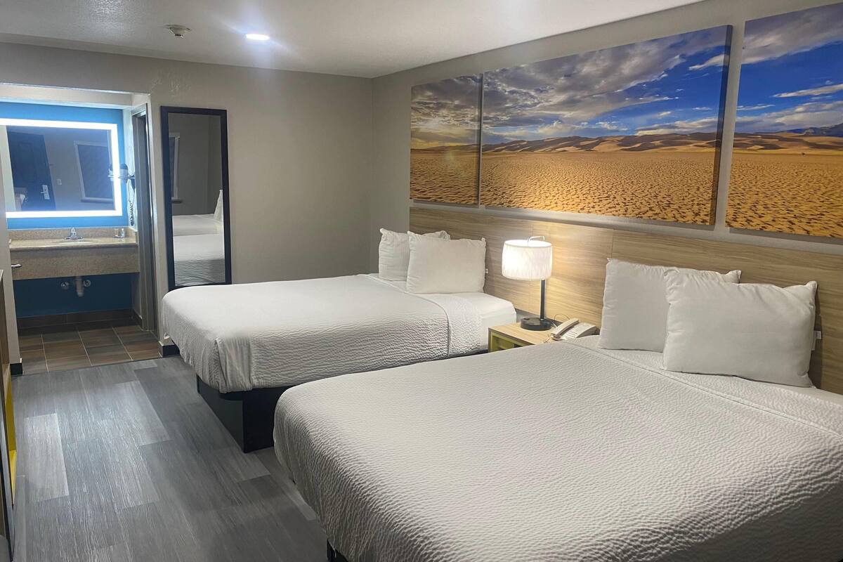 A hotel room with two beds, a bathroom with a mirror and sink, and a desert landscape painting on the wall.