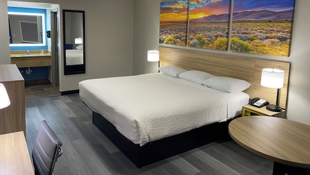 A hotel room with a bed, bedside lamps, a round table, a mirror, and a painting of a sunset over a desert landscape. - Days Inn by Wyndham El Paso West (El Paso)