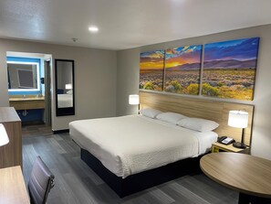 A hotel room with a bed, bedside lamps, a round table, a mirror, and a painting of a sunset over a desert landscape.