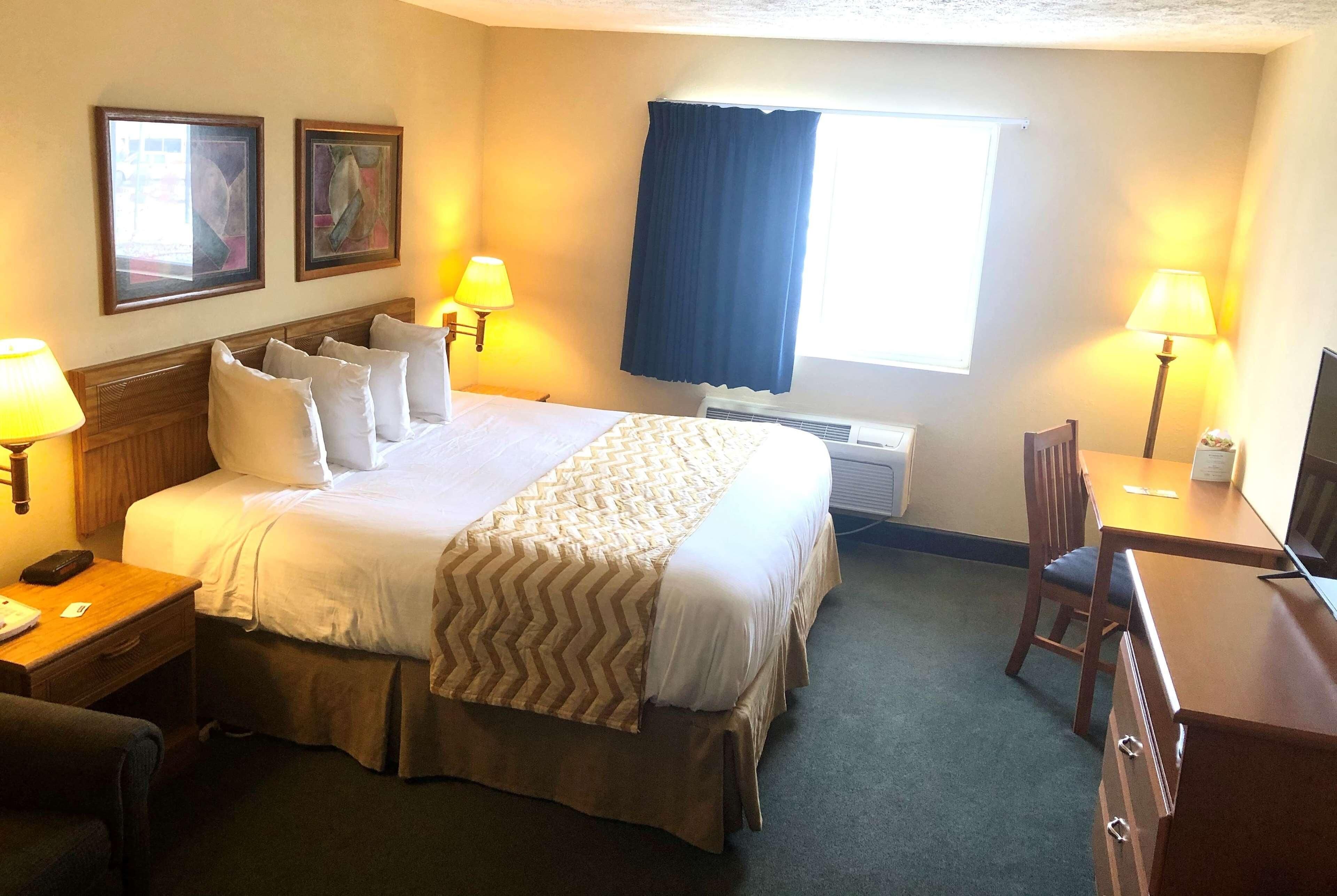 A hotel room with a bed, bedside tables, a desk, a chair, a window with curtains, and wall-mounted artwork.