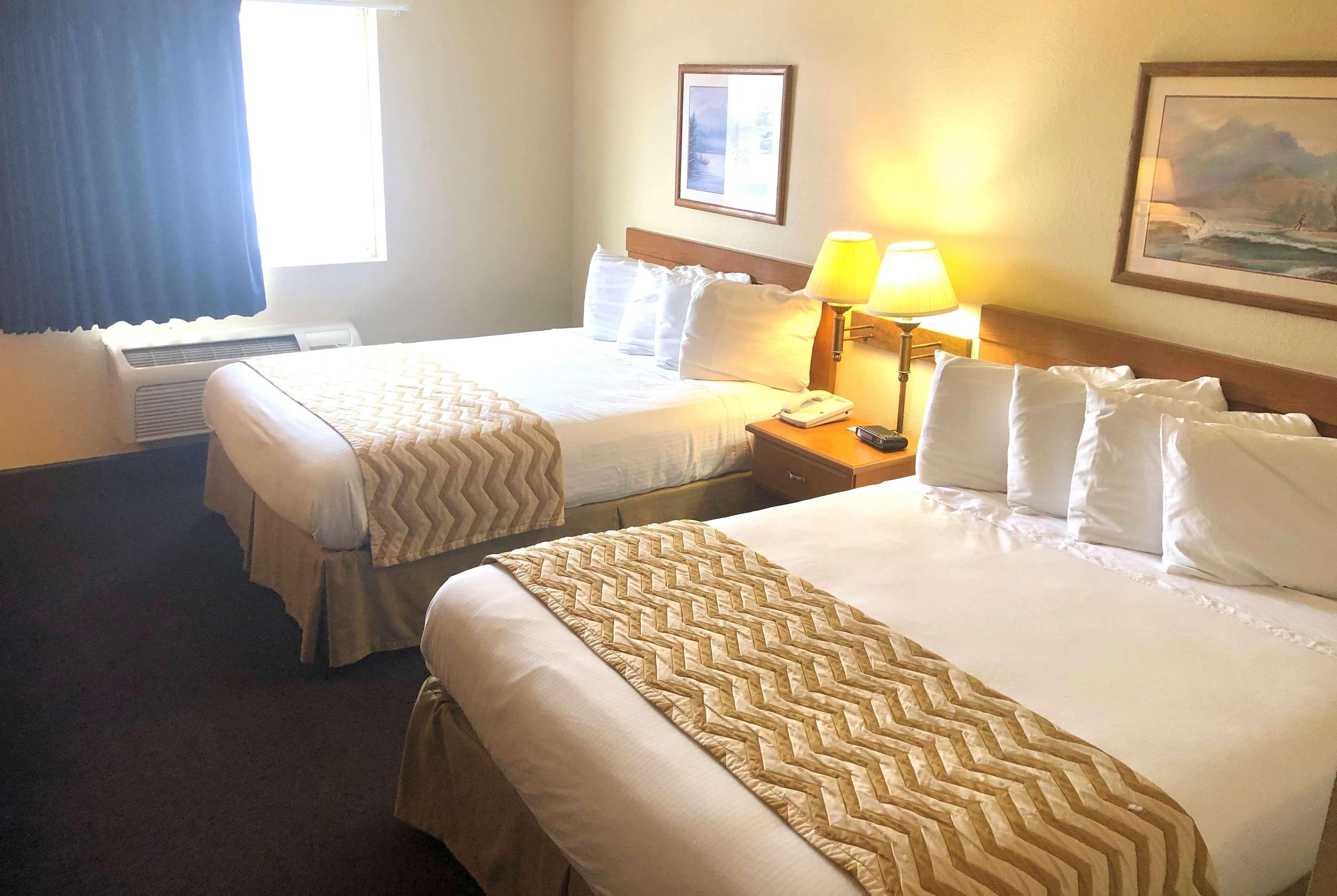 A hotel room with two beds, a bedside table with a lamp, a framed picture on the wall, and an air conditioning unit.