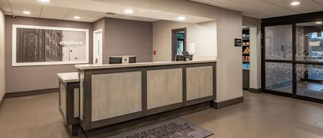 A reception area with a counter, a framed picture on the wall, and a doormat with a message.