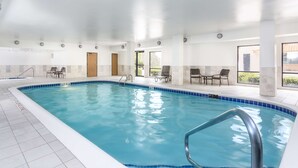 Una piscina interior con agua cristalina, paredes con azulejos y algunas sillas y mesas cerca de las ventanas.