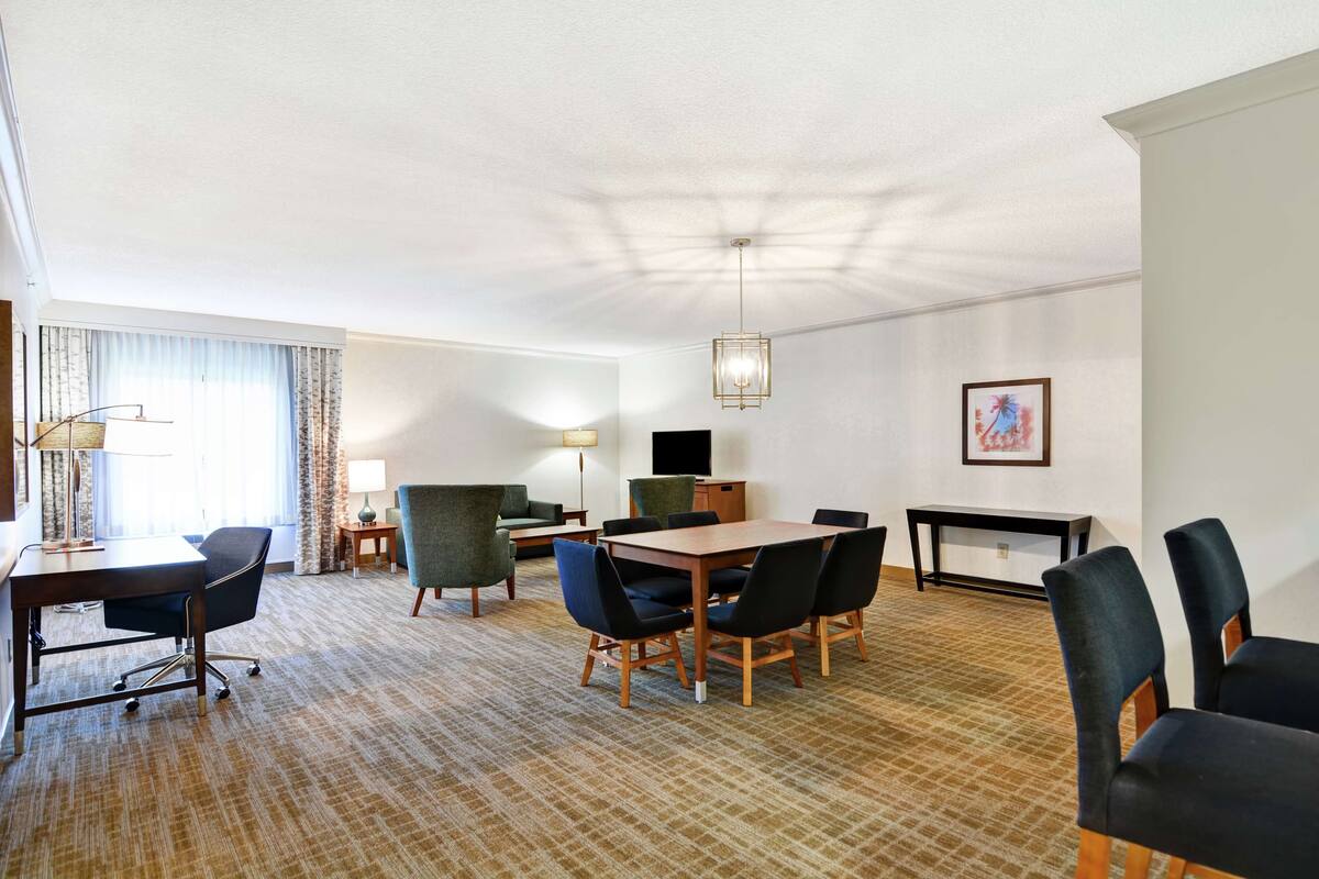 Quarto de hotel com uma cama grande, uma mesa de escritório com cadeira, uma mesa de jantar com cadeiras, uma televisão e um quadro pendurado na parede.
