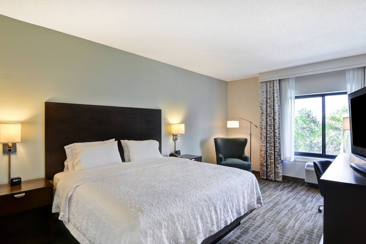 Quarto de hotel com uma cama grande, duas luminárias de cabeceira, uma cadeira, uma escrivaninha com televisão e uma janela com cortinas.