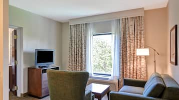 Une chambre d’hôtel avec un fauteuil vert, un canapé, une petite table, un téléviseur posé sur un meuble en bois et une fenêtre avec des rideaux.