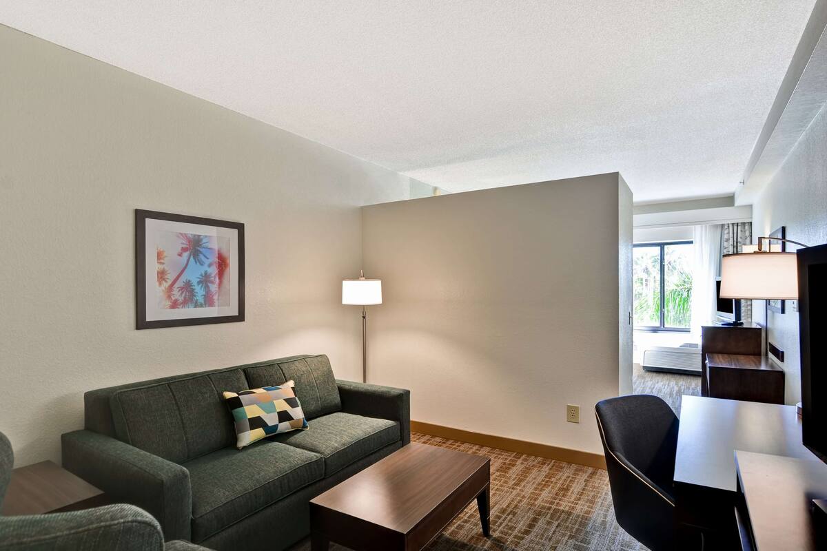Quarto de hotel com sofá verde, mesa de centro de madeira, luminária, quadro na parede e vista do exterior pela janela.