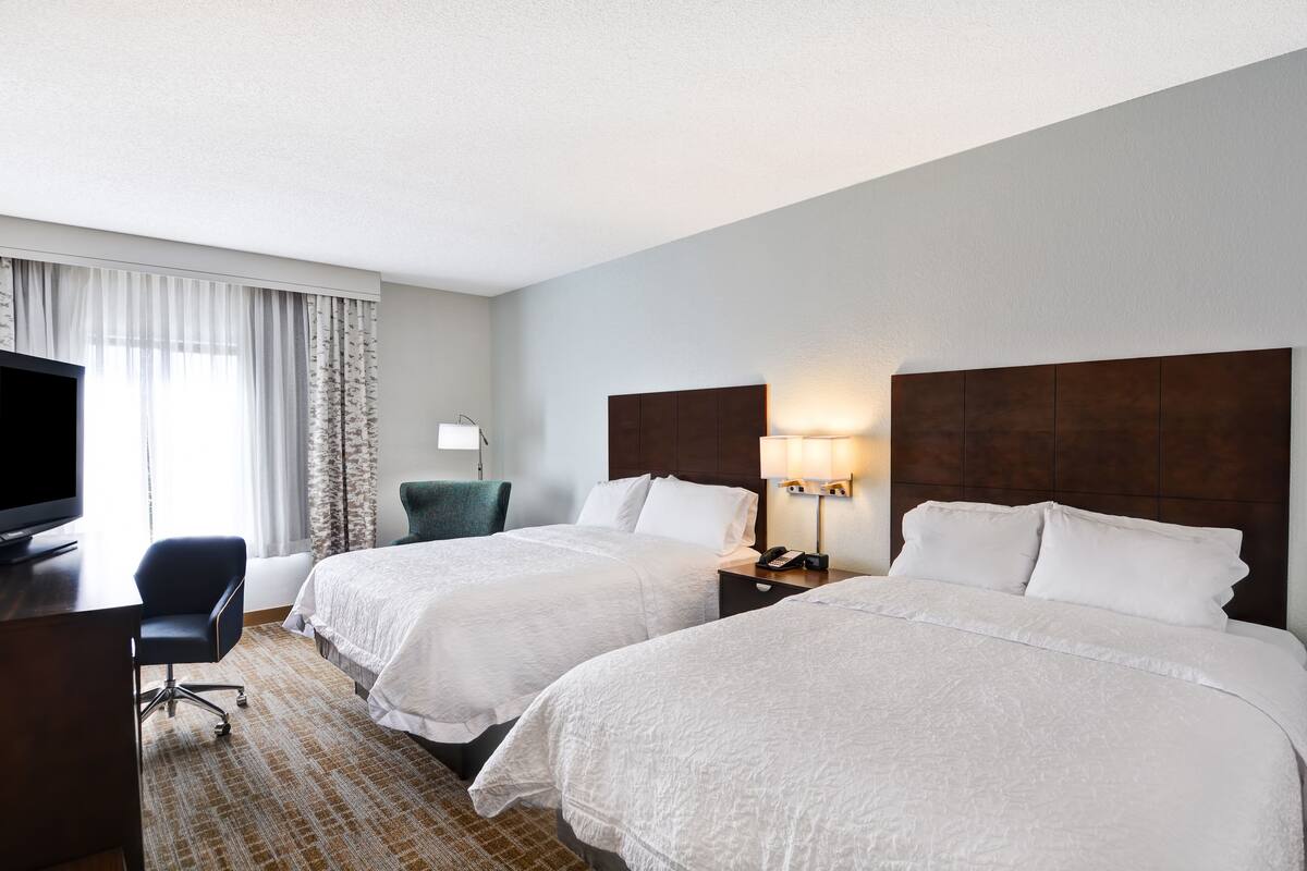 Quarto de hotel com duas camas, uma escrivaninha com televisão, uma cadeira e um abajur.