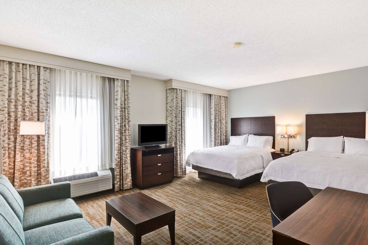 Quarto de hotel com duas camas, uma televisão, uma escrivaninha e um sofá.