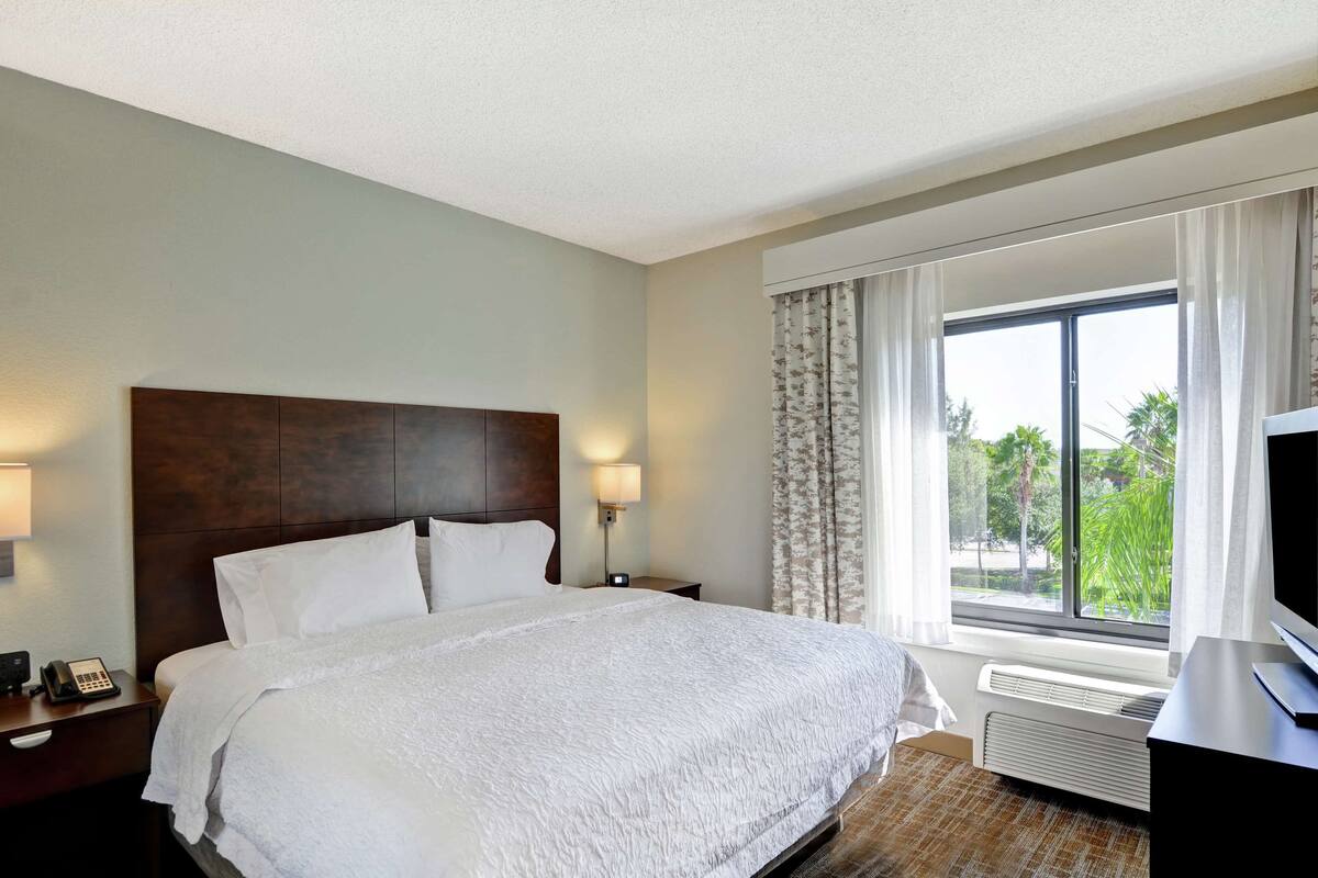 Quarto de hotel com uma cama grande, televisão, mesa de cabeceira com telefone e janela com cortinas.