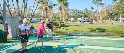 A shuffleboard court with players, palm trees, and RVs in the background.