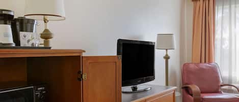 Habitación de hotel con televisión, cafetera, microondas, escritorio de madera, una silla rosa y una ventana con cortinas.