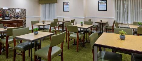 A hotel dining area with multiple tables and chairs, a kitchen area, and framed pictures on the walls.