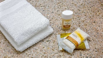 Una pila de toallas blancas, una botella de Champú Hidratante Pantene y un tubo de Gel de Ducha Revitalizante Fresh Clean sobre una encimera de granito moteado.