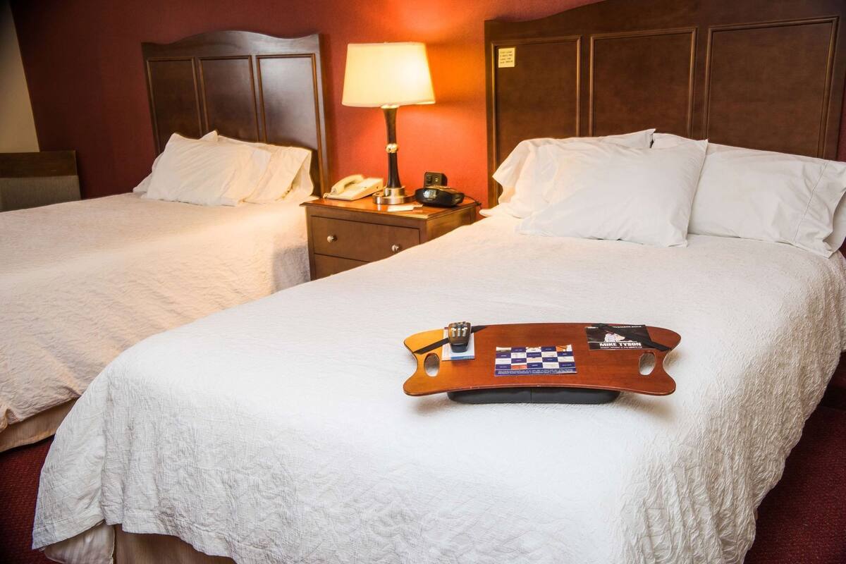 A hotel room with two beds, a nightstand with a lamp, a telephone, and a tray with a chessboard and remote controls.