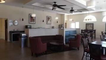 A spacious living area with a curved sofa, a dining table, and chairs. There are ceiling fans, pendant lights, and a kitchen area in the background.