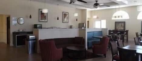 A spacious living area with a curved sofa, a dining table, and chairs. There are ceiling fans, pendant lights, and a kitchen area in the background.