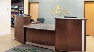 A reception desk with a wooden finish and a marble countertop, a sign above it reading "Wingate by Wyndham", and a doormat with the same branding.