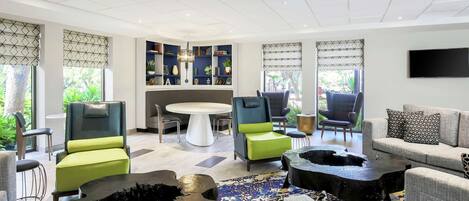 A modern living room with a round table, green and grey upholstered chairs, a sofa, and a large TV on the wall.