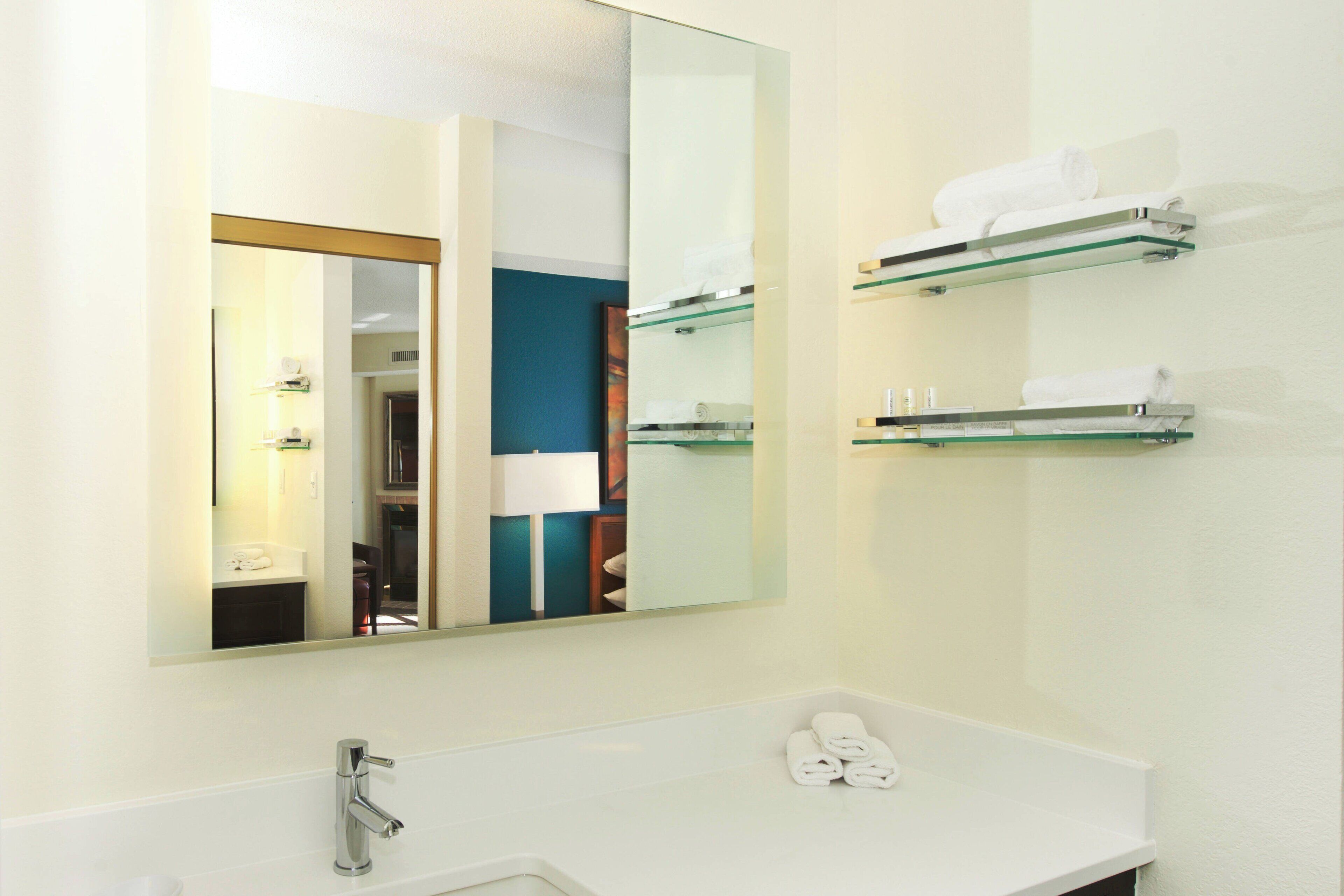 A modern bathroom with a large mirror, a white sink, and glass shelves holding towels and toiletries.