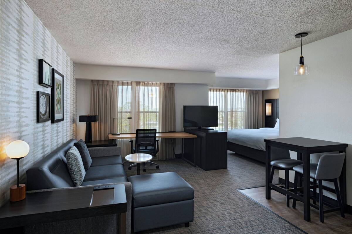 A modern hotel room with a sofa, desk, TV, and dining area.