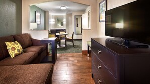 Hotelkamer met een bruine bank, een televisie op een dressoir, een zithoek met een tafel en stoelen, en een keuken op de achtergrond.