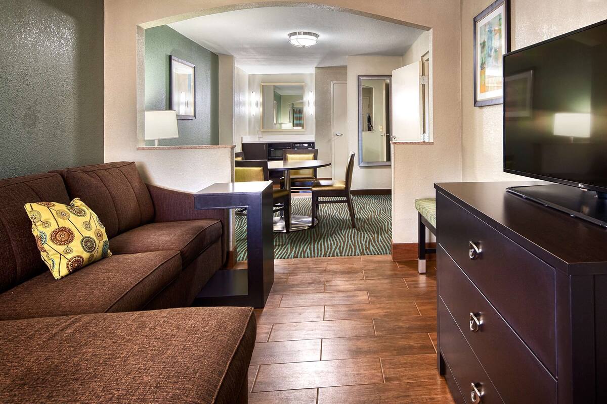 A hotel room with a brown sofa, a television on a dresser, a dining area with a table and chairs, and a kitchen in the background.