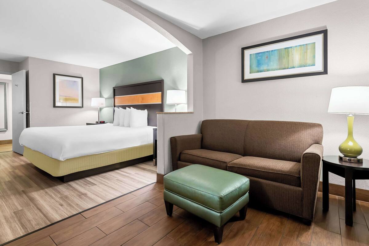A hotel room with a bed, a sofa, an ottoman, a lamp, and a painting on the wall.
