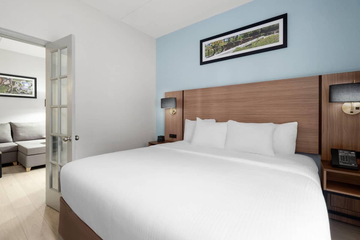 A hotel room with a large bed, a wooden headboard, a bedside table with a lamp, a small desk, and a chair.