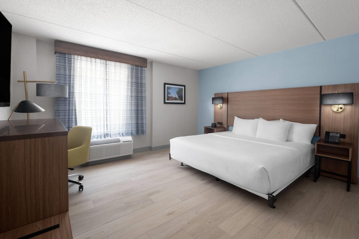A hotel room with a large bed, a desk with a lamp, a chair, a bedside table with a phone, and a window with curtains.