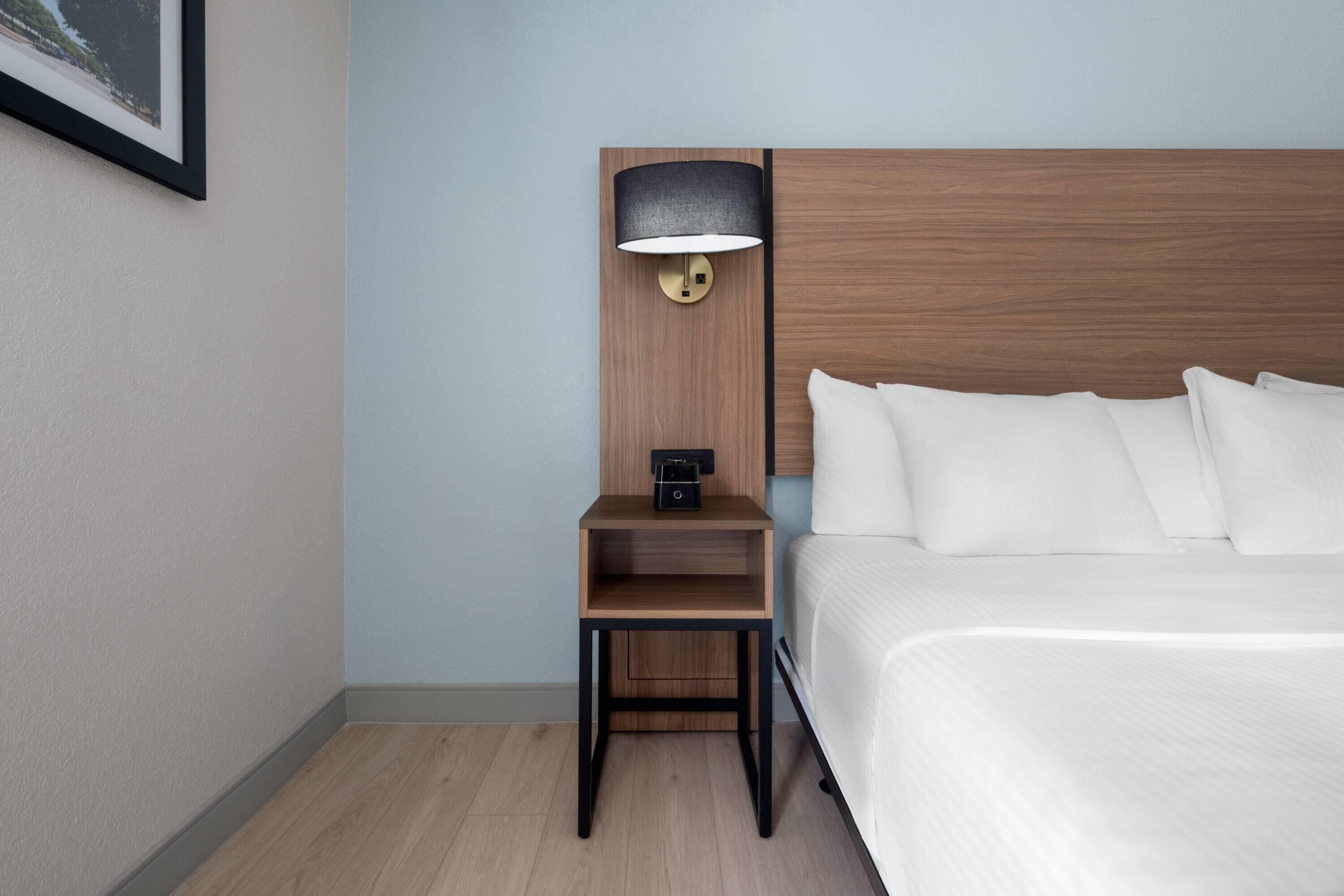 A neatly arranged hotel room with a bed, a nightstand with a lamp, and a small shelf.