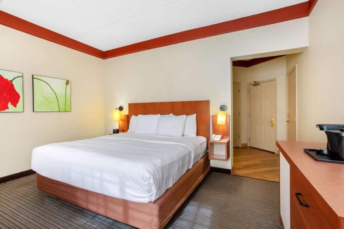 A hotel room with a bed, bedside lamps, a desk, and a door leading to another room.