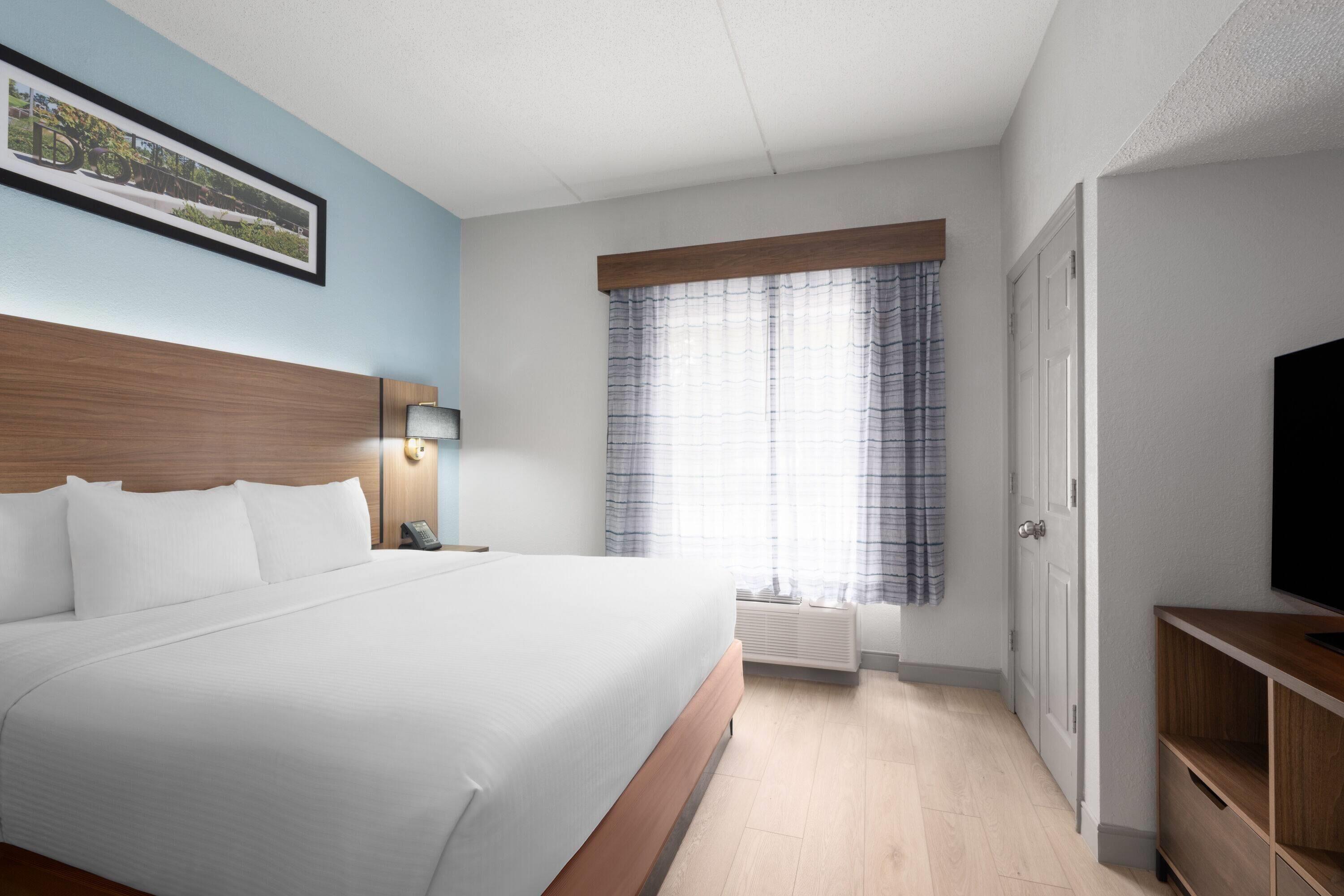 A hotel room with a large bed, a bedside table with a lamp, a window with curtains, and a television on a wooden stand.