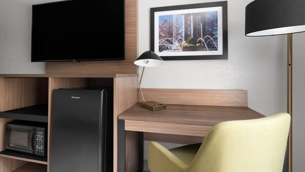 Une chambre d’hôtel moderne équipée d’un téléviseur à écran plat fixé au mur, d’un tableau représentant une fontaine, d’un bureau avec une lampe, d’un réfrigérateur Hisense et d’un four à micro-ondes.