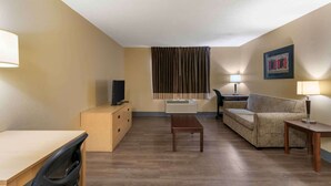 A compact hotel room with a wooden floor, a sofa, a small table, a TV on a dresser, and a window with curtains. - Extended Stay America Suites Kansas City Overland Park Quivi (Overland Park)