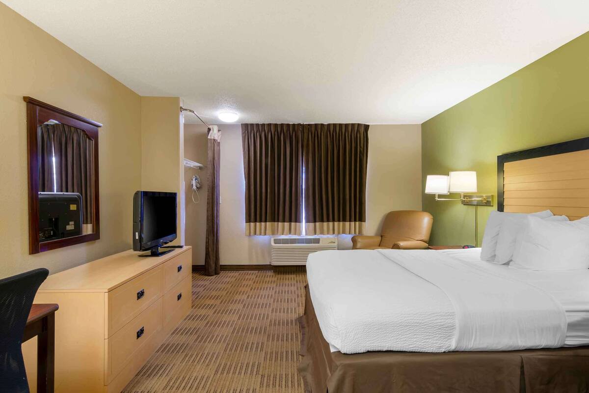 A hotel room with a bed, a desk with a television, a chair, a window with curtains, and a wall-mounted lamp.