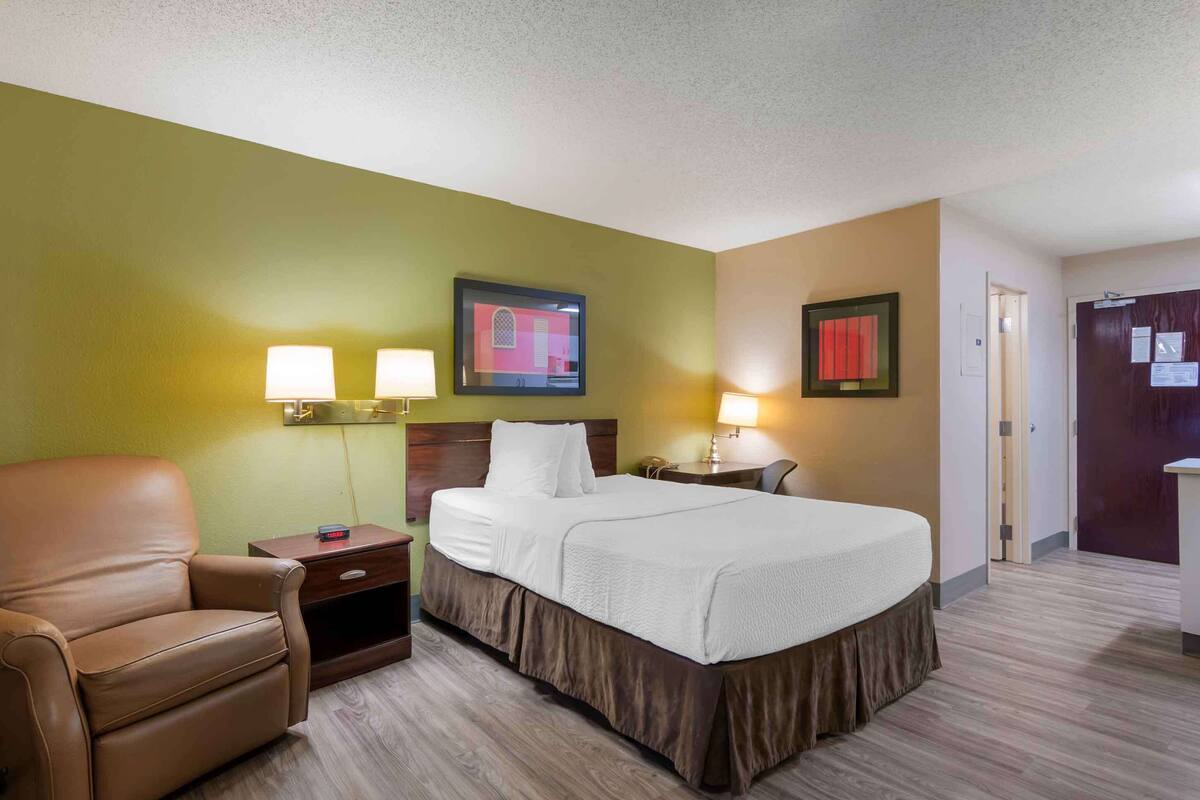 A hotel room with a bed, a chair, two bedside lamps, and a framed picture on the wall.