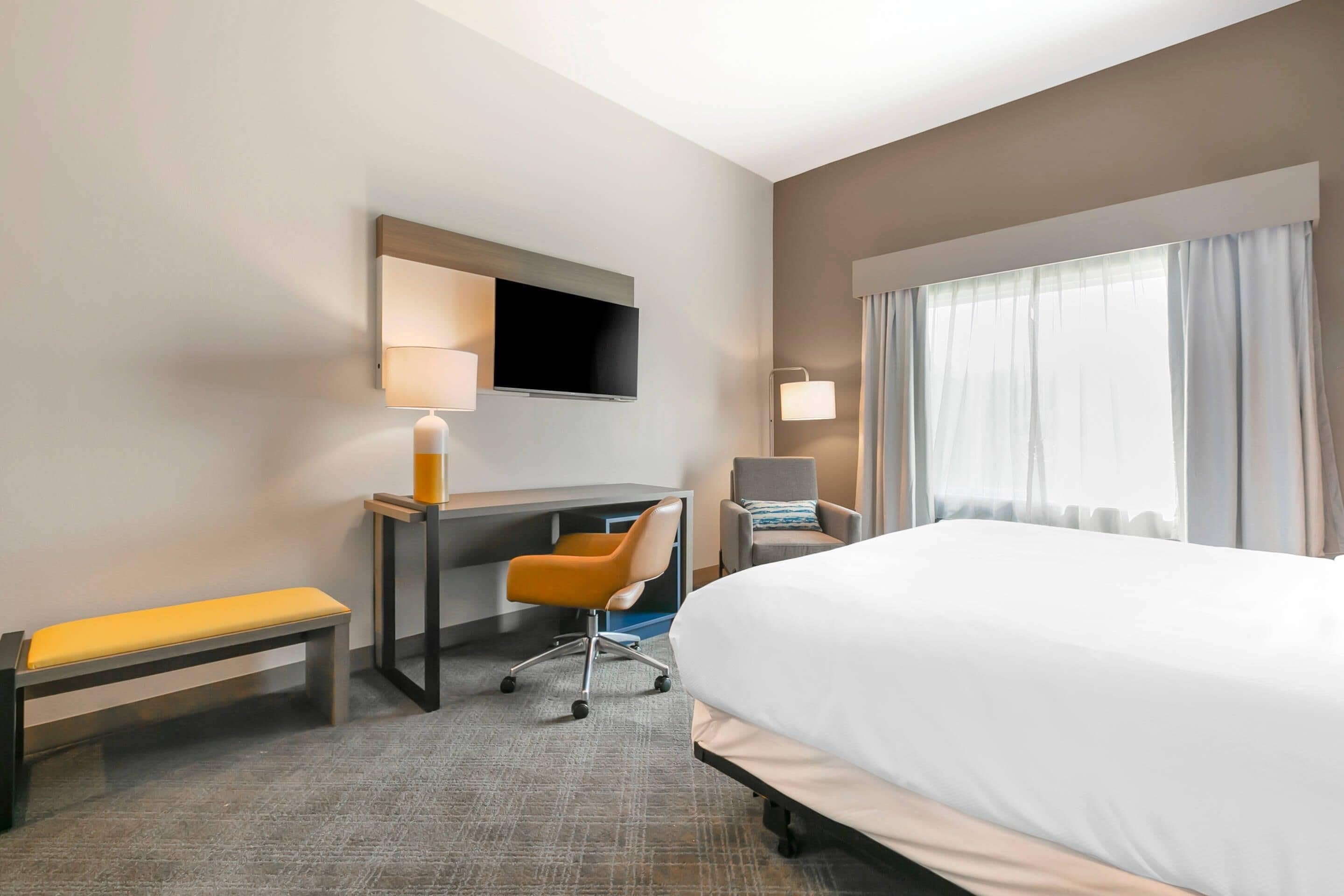 A hotel room with a large bed, a desk with a chair, a bench, a TV mounted on the wall, and a window with curtains.
